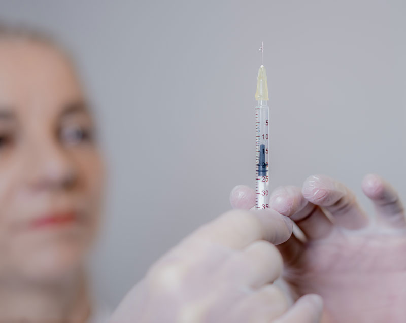 Vorbereitung einer Botox-Injektion in der Praxis Nina Bettge, Fokus auf eine präzise gefüllte Spritze mit feiner Nadel im Vordergrund, unscharfe Darstellung einer erwartungsvollen Patientin im Hintergrund.