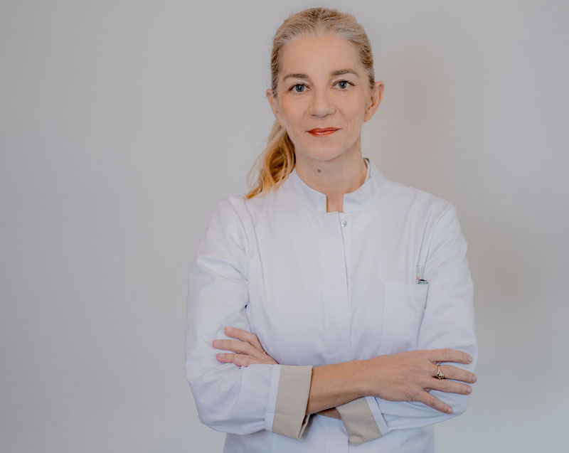Kompetente Medizinerin Nina Bettge in professioneller weißer Arztkleidung, steht selbstbewusst mit verschränkten Armen vor einem neutralen Hintergrund, strahlt Vertrauenswürdigkeit und Fachkenntnis aus.